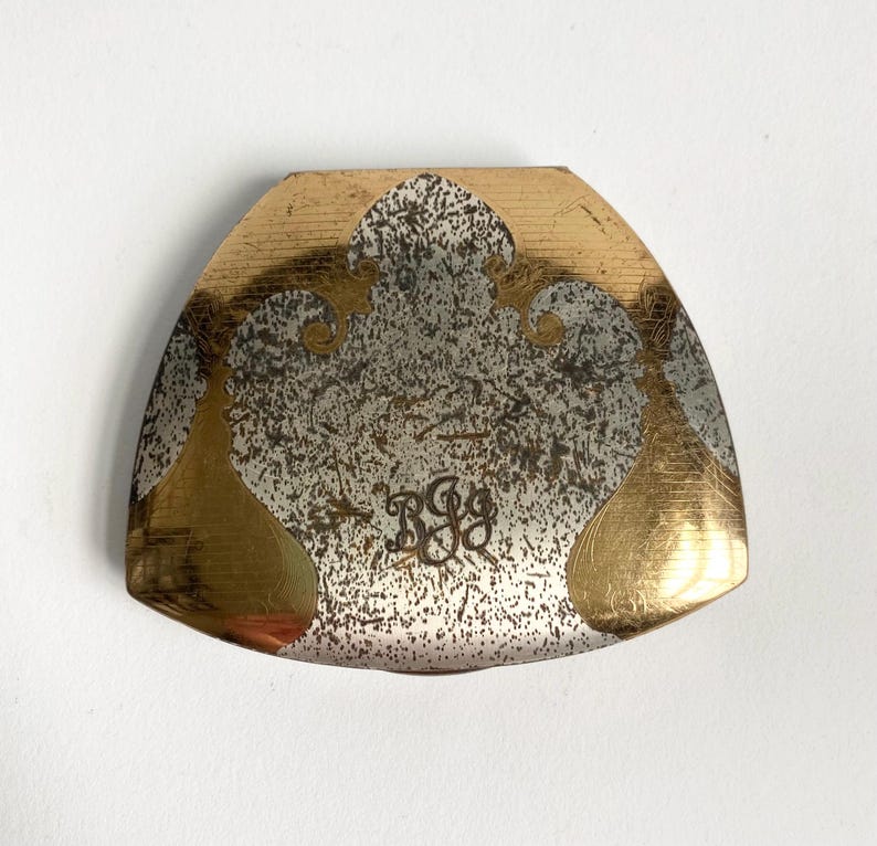 May include: A gold and silver-toned metal object with a curved, shield-like shape. The top and edges are gold, while the center features a textured silver design with the initials "RGG" in a decorative script. The object has a vintage aesthetic.