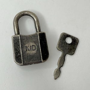 May include: A small, silver-toned padlock with a matching key. The padlock has the word "KID" printed on the front. The key is rectangular with a circular hole at the top.