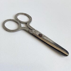 May include: Vintage silver-toned scissors with ornate detailing on the finger loops and the hinge area. The blades are a contrasting silver color. The scissors are marked "STEEL USA".
