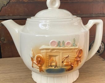 May include: A white ceramic teapot with a brown and orange scene of a fireplace and a spinning wheel. The teapot has a lid with a knob on top.