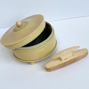 May include: An antique ivory-colored round box with a lid, and a small, oval-shaped object. The box has a dark interior and rests on three small feet. The lid is slightly ajar, revealing the interior. The oval object is light-colored with a darker base.