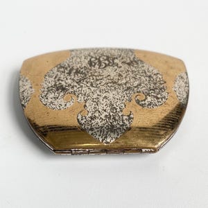 May include: A vintage compact mirror with a gold-toned frame and a decorative silver and gold design on the lid. The lid features an ornate pattern with a monogram in the center. The compact is closed and appears to be in good condition.