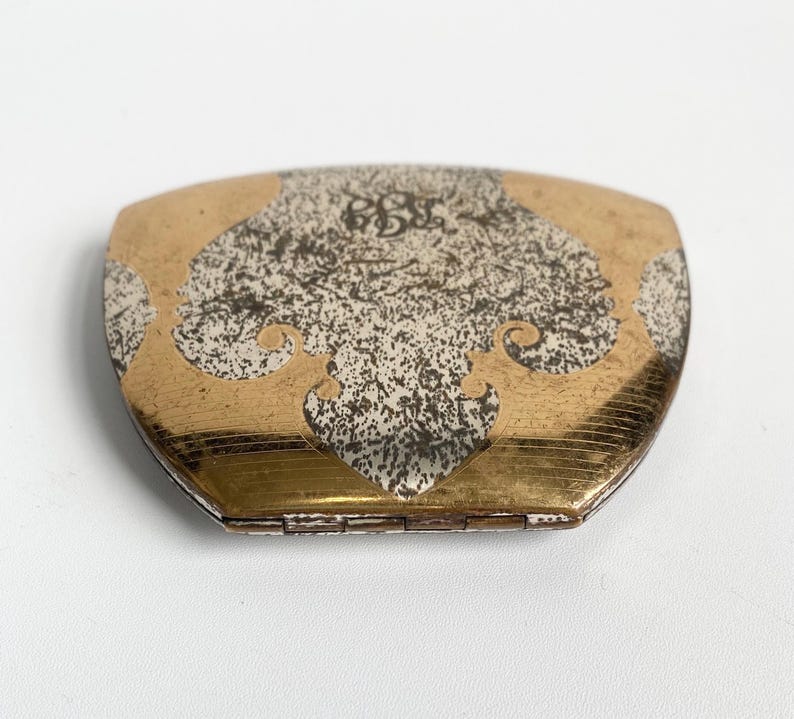 May include: A vintage compact mirror with a gold-toned frame and a decorative silver and gold design on the lid. The lid features an ornate pattern with a monogram in the center. The compact is closed and appears to be in good condition.