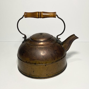 May include: Antique copper kettle with a wooden handle. The kettle has a rounded body, a spout, and a lid. The handle is made of light brown wood. The kettle is a dark copper color with some discoloration.