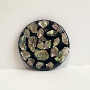 May include: A round, black decorative object inlaid with iridescent abalone shell pieces. The shell fragments vary in size and shape, displaying a range of colors including silver, green, and pink. The object is set against a white background.