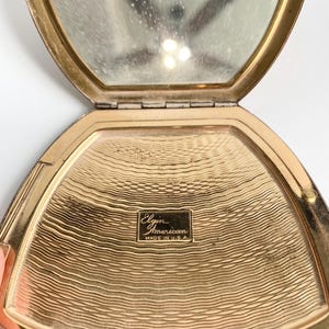 May include: Open vintage gold-toned compact mirror with a hinged lid. The interior features a mirror and a textured gold surface. The compact is marked "Elgin American MADE IN U.S.A."