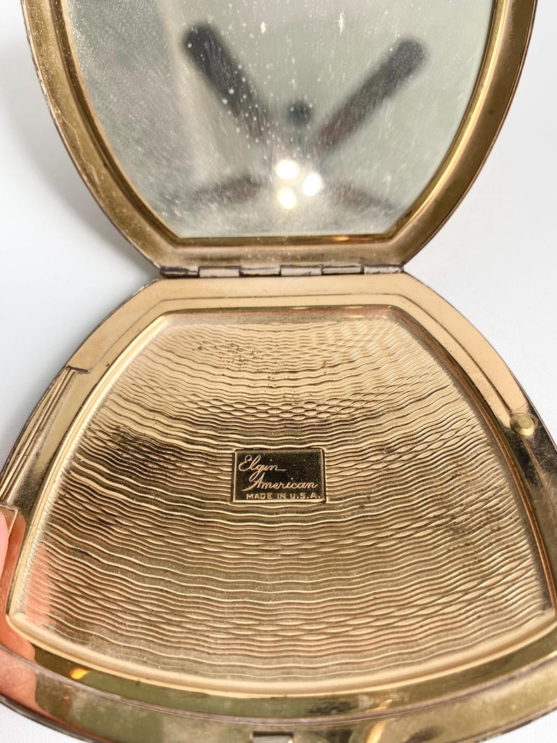 May include: Open vintage gold-toned compact mirror with a hinged lid. The interior features a mirror and a textured gold surface. The compact is marked "Elgin American MADE IN U.S.A."