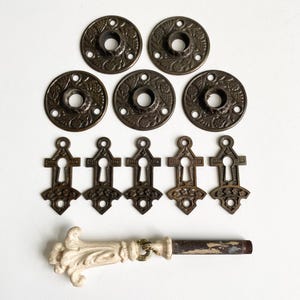 May include: An assortment of antique hardware components, including six circular backplates with floral designs, six keyhole covers, and a decorative door handle with a white porcelain grip and metal shaft. The hardware has a vintage, aged appearance.