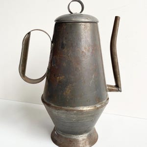May include: A vintage metal teapot with a dark, aged patina. The teapot features a curved spout, a handle, and a lid with a circular handle. The body has a conical shape, and the base is slightly flared. The overall aesthetic suggests a rustic, antique style.