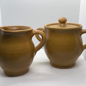 May include: A set of two brown ceramic kitchenware pieces, a creamer and a sugar bowl with a lid.
