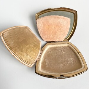 May include: Open vintage gold-toned compact with a mirror, powder, and a peach-colored powder puff. The compact is open, revealing the powder and puff. The lid has a textured design. The compact is on a white surface.