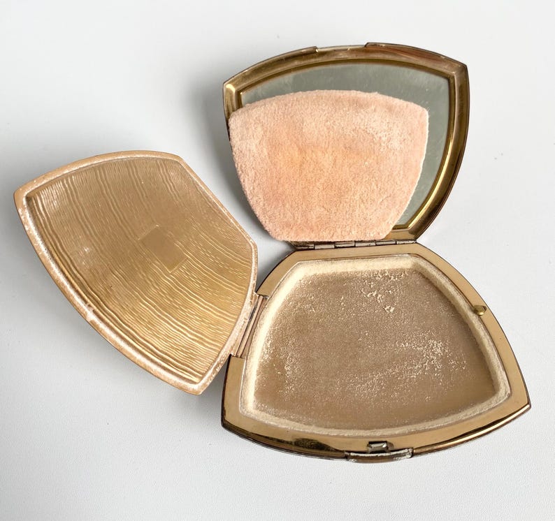 May include: Open vintage gold-toned compact with a mirror, powder, and a peach-colored powder puff. The compact is open, revealing the powder and puff. The lid has a textured design. The compact is on a white surface.