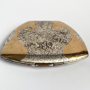 May include: A vintage, silver-toned compact with gold-colored accents. The compact has a unique shape and features an ornate design on the top. The design includes a monogram in the center.