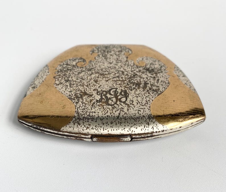 May include: A vintage, silver-toned compact with gold-colored accents. The compact has a unique shape and features an ornate design on the top. The design includes a monogram in the center.