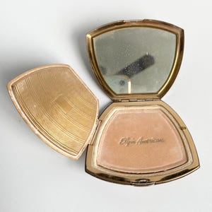 May include: Open vintage gold-toned compact with a mirror and powder. The compact is open, revealing a light pink powder and the engraved text "Elgin American". The lid has a decorative pattern.