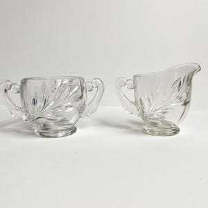 May include: Clear glass sugar bowl with two handles and a matching creamer. Both pieces feature a leaf design etched into the glass. The sugar bowl has a round shape, while the creamer has a spout for pouring.