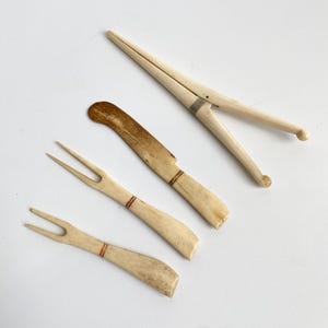 May include: An assortment of antique ivory-colored serving utensils. The set includes a nutcracker, a knife, and two forks. The knife and forks have a reddish-brown band near the handle. The nutcracker has a silver-colored band.