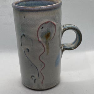 May include: A white ceramic mug with a light blue glaze and a pink and brown balloon design. The mug has a handle and is decorated with a pink and brown balloon with a string.