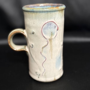 May include: Handmade ceramic mug with a whimsical balloon design. The off-white mug features a blue rim and handle, with a hand-painted balloon illustration in shades of blue and purple. The mug is approximately 5 inches tall.