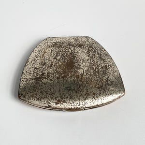 May include: A vintage, silver-toned compact mirror with a unique, semi-circular shape. The surface has a distressed, speckled appearance with a darker, central area. The compact's edges are smooth, and the overall design suggests an antique or retro aesthetic.