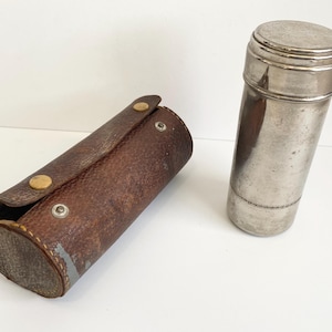 May include: A vintage silver metal container with a lid next to a brown leather case. The cylindrical container has a shiny finish. The leather case is cylindrical with a snap closure and is brown with a textured surface.
