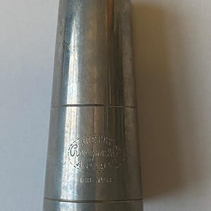 May include: A silver metal clarinet mouthpiece with the inscription "Buffet Crampon & Cie A Paris France".
