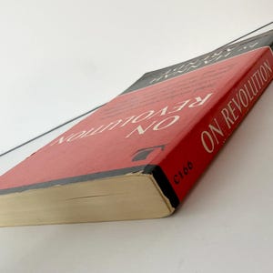 May include: A paperback book titled On Revolution by Hannah Arendt with a red cover, black borders, and white text. The book is positioned at an angle, showing the front cover and the spine, which features the author's name and the title in white.