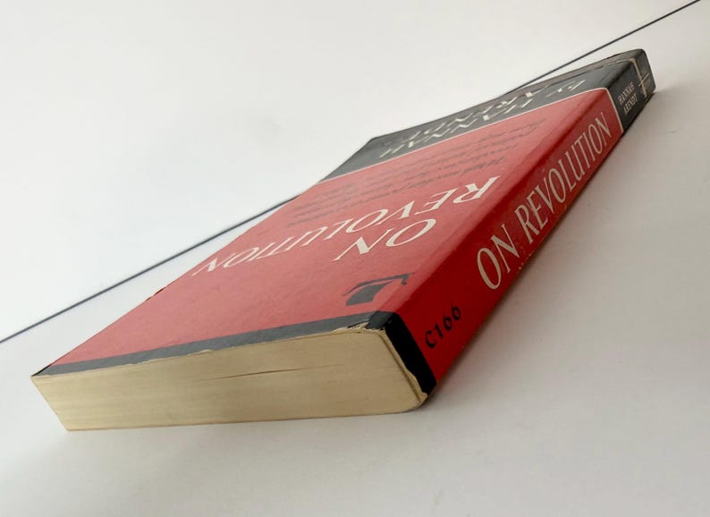 May include: A paperback book titled On Revolution by Hannah Arendt with a red cover, black borders, and white text. The book is positioned at an angle, showing the front cover and the spine, which features the author's name and the title in white.