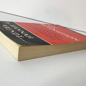 May include: A paperback book titled On Revolution by Hannah Arendt. The cover features a red upper section and a black lower section with white text. The book is positioned at an angle, showing the front cover and the off-white page edges.