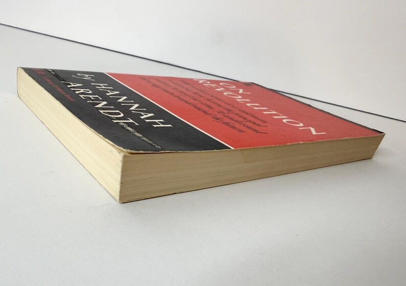 May include: A paperback book titled On Revolution by Hannah Arendt. The cover features a red upper section and a black lower section with white text. The book is positioned at an angle, showing the front cover and the off-white page edges.