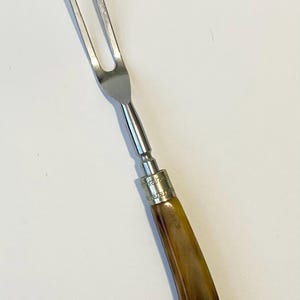 May include: A vintage carving fork with a stainless steel head and a curved handle. The handle is a warm brown color with a glossy finish. The fork has two tines and a decorative metal band.