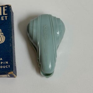 May include: A vintage, light blue Kent hair clip with a ribbed design. The clip has the brand name "KENT" embossed on its surface. A partially visible blue box is on the left side, with the text "PIN PRODUCT".