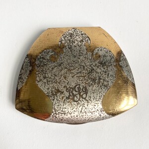 May include: A vintage, triangular-shaped metal object with a gold and silver finish. The design features gold accents and a speckled silver center, with the initials "BGG" engraved in the center. The edges are rounded.