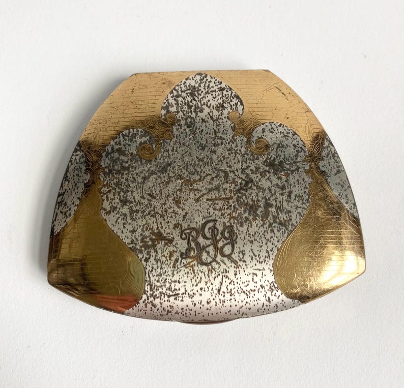 May include: A vintage, triangular-shaped metal object with a gold and silver finish. The design features gold accents and a speckled silver center, with the initials "BGG" engraved in the center. The edges are rounded.