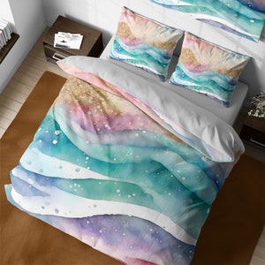 May include: A white duvet cover with a watercolor design of turquoise, pink, and purple waves with gold glitter accents. The duvet cover is on a bed with two matching pillowcases.