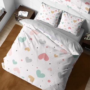 Op de afbeelding: Wit beddengoed met een patroon van kleurrijke harten en stippen. De beddengoedset bevat een dekbedovertrek en twee kussenslopen. Het dekbedovertrek heeft een websiteadres erop gedrukt: https://cozywhimsicaletsy.com