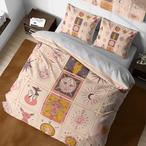 May include: A cream-colored duvet cover with a pink and gold geometric pattern featuring suns, moons, stars, and other celestial symbols. The duvet cover is on a bed with two matching pillowcases.