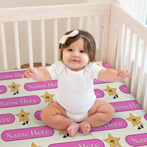 May include: A pink and cream baby crib sheet with a repeating pattern of yellow stars with smiling faces and the text "Name Here". A baby is sitting on the sheet.