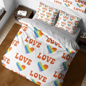 May include: White bedding featuring a rainbow heart and the word "LOVE" repeated in a retro font. The bedding is displayed on a bed with two matching pillowcases.