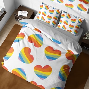 May include: White bedding with a rainbow heart pattern. The bedding includes a duvet cover and two pillowcases. The duvet cover has a white background with rainbow hearts scattered across the surface. The pillowcases have a white background with rainbow hearts arranged in a repeating pattern.