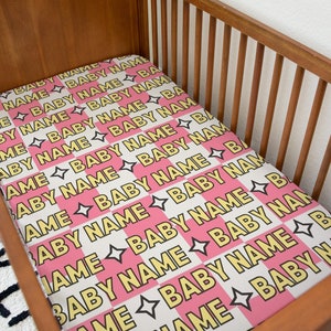 May include: A pink and white checkered crib sheet with the words "Baby Name" repeated in yellow.