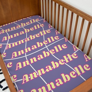 May include: A purple crib sheet with a white spiderweb pattern and the name "Annabelle" repeated in yellow and pink letters.