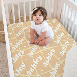 May include: A baby in a white onesie sits on a crib sheet. The sheet is tan with a grid pattern and the name "Ashley" repeated in white. The crib is made of light-colored wood.