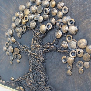 Puede incluir: Una obra de arte enmarcada que representa un árbol hecho de botones dorados sobre un lienzo gris oscuro. El tronco del árbol está hecho de hilo marrón.