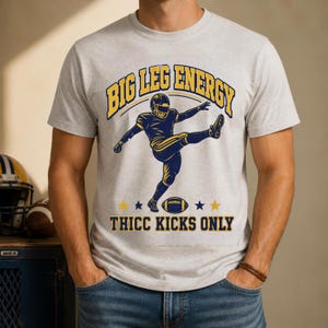 May include: A light grey crew neck t-shirt featuring a graphic of a football player kicking a ball. The text "BIG LEG ENERGY" appears in yellow and blue above the player, and "THICC KICKS ONLY" is printed in blue below the football graphic.