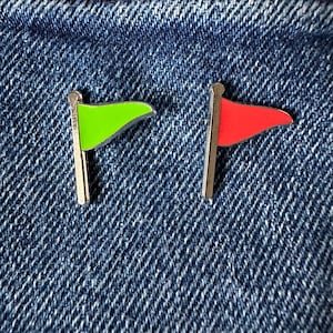 Könnte beinhalten: Zwei kleine Flaggen-Anstecknadeln, eine grüne und eine rote, vor einem blauen Jeans-Hintergrund. Jede Nadel hat einen silberfarbenen Stab und eine dreieckige Flaggenform. Die Nadeln sind wahrscheinlich Accessoires oder Dekorationsartikel.