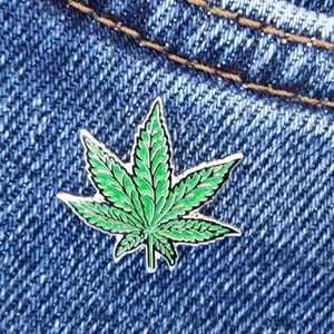 Op de afbeelding: Een groene marihuana-blad emaille pin op een blauwe denim achtergrond.