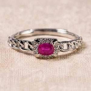 Könnte beinhalten: Ein silberfarbener Ring mit einem zentralen, oval geschliffenen, leuchtend pink-roten Edelstein. Der Stein ist von einem Kranz aus kleinen, klaren Steinen umgeben. Die Ringschiene ist als polierte Metall-Gliederkette gestaltet.