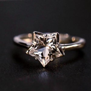 Könnte beinhalten: Ein silberfarbener Ring mit einem markanten, sternförmigen, klaren Edelstein in der Mitte. Die Ringschiene ist glatt und poliert und hält den facettierten Sternstein sicher mit mehreren kleinen Metallkrappen, was für ein helles Aussehen sorgt.
