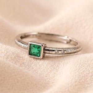 Könnte beinhalten: Ein silberner Ring mit einem quadratischen, smaragdgrünen Edelstein. Das Band hat ein strukturiertes Design mit kleinen, klaren Akzentsteinen. Der Ring liegt auf einem weichen, cremefarbenen Hintergrund. Ein schlichtes Schmuckstück.